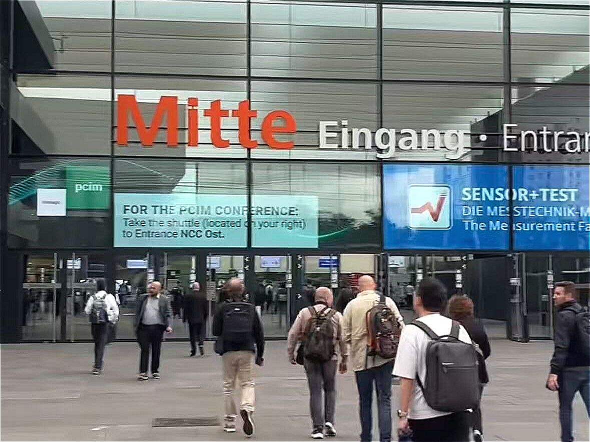 Modern glass building entrance with 'Mitte Eingang' signage; people walking toward conference entrances.